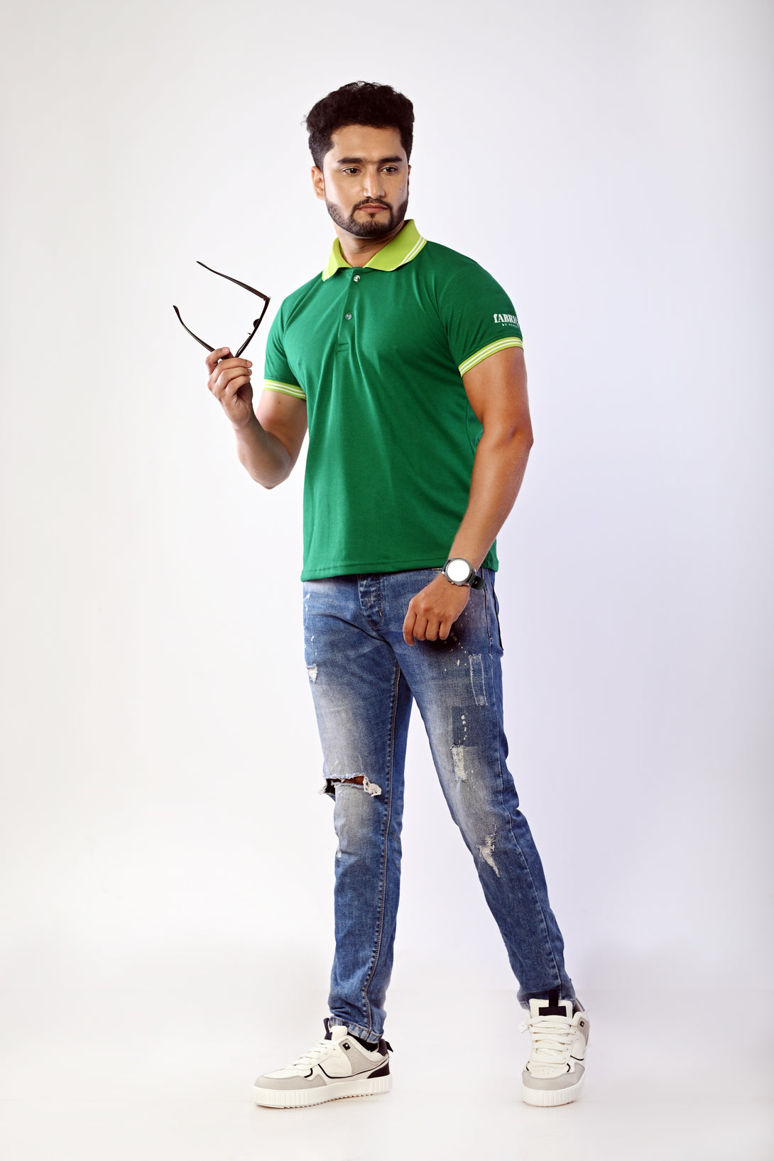 Man wearing a green polo shirt and blue jeans holding sunglasses on a white background