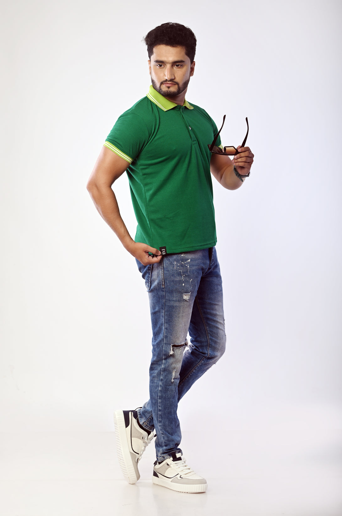 Man wearing a green polo shirt and blue jeans holding sunglasses on a white background